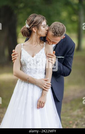 Portrait of amazing wedding couple standing in park in summer. Young beautiful woman bride kissing temple of man. Love. Stock Photo