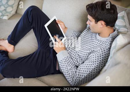 Where do i switch this thing on. a young man lying on his sofa while using his digital tablet. Stock Photo