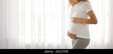 Expectant mother gently strokes and hugs her pregnant tummy while standing against bright window. Stock Photo