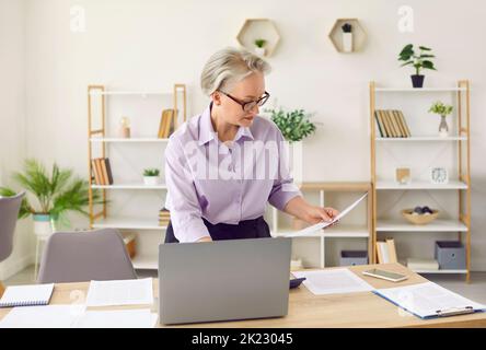 Serious mature businesswoman working in her office, doing paperwork and using laptop Stock Photo