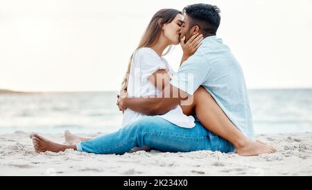 Kiss, beach and love of couple on a date for anniversary, valentines day or romance summer holiday with clear sky, ocean waves and sand. Romantic Stock Photo