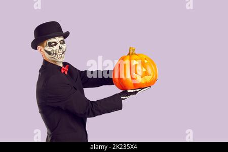 Cheerful man with traditional Halloween skeleton make-up holding glowing pumpkin in his palm. Stock Photo