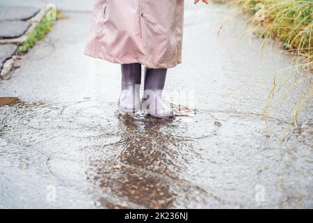 Little girl in pink waterproof raincoat, purple rubber boots funny jumps through puddles on street road in rainy day weather. Spring, autumn. Children Stock Photo