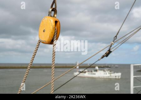 Close-up of a rope pulley and ropes on an old sailing ship against a landscape with sea, clouds and a ship. Stock Photo