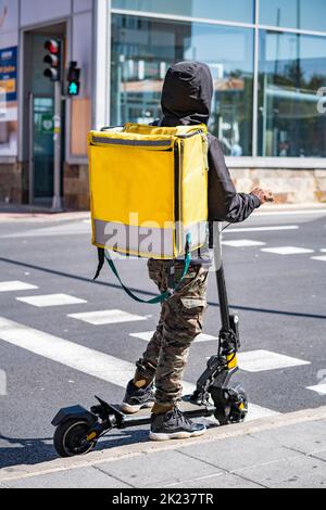 Man courier online food delivery with yellow thermal backpack waiting the green traffic light on an electric scooter. Stock Photo