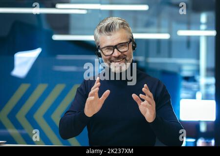 Senior gray haired businessman talking on video call, using headset investor in glasses smiling and looking at camera, webcam view man working inside modern office building. Stock Photo