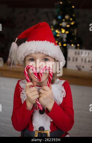 cute child in red santa hat, holding heart-shaped lollipops in front of his face. kid is waiting for a miracle on Christmas Eve. New Year, merry winte Stock Photo