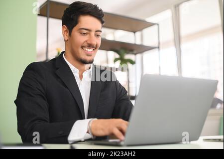 Smiling optimistic young latin entrepreneur wearing smart casual clothes, sitting at the desk, typing on keyboard, businessman professional is feeling ready to work and start new projects Stock Photo