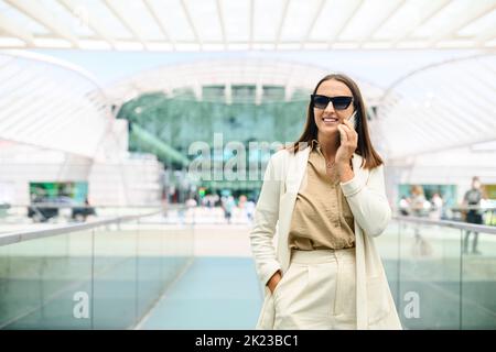 Smiling successful contemporary businesswoman manager in smart casual wear holding phone call. Inspired caucasian female small business owner talking on the smartphone walking outdoors Stock Photo