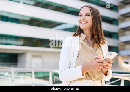 Young woman in stylish smart casual suit standing in the city downtown and holding smartphone. Female spending time online, texting, scrolling news feed in social networks, using online map Stock Photo