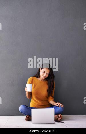 Feeling bright and perky with the help of coffee. A young woman drinking coffee while sitting on the floor with her laptop and mobile phone. Stock Photo