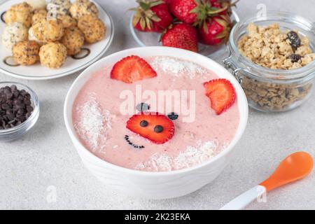Fun food for kids - smoothie-bowl in shape funny piglet, with strawberries, granola, chocolate and crispy balls on light gray background Stock Photo