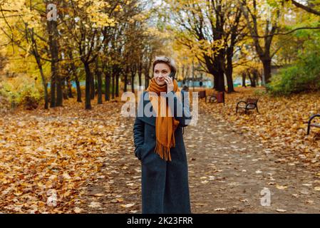 A blonde in a coat and scarf talks on the phone as she walks through an autumn park. Stock Photo