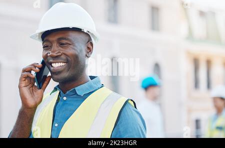 Construction worker using a smartphone for phone call on site. Portrait of happy, smiling and black businessman in safety helmet and safety gear on Stock Photo