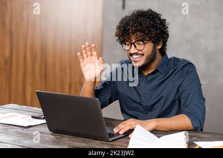 Video call concept, virtual meeting. Young middle eastern guy relaxed sitting at the table and talking online via video call with group of friends or employees. Man waving to the camera Stock Photo