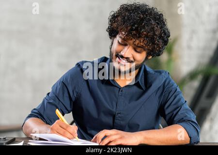 Online training. Young guy student with curly hair studying at home while sitting in the modern room. He writing with a pen in a notebook Stock Photo