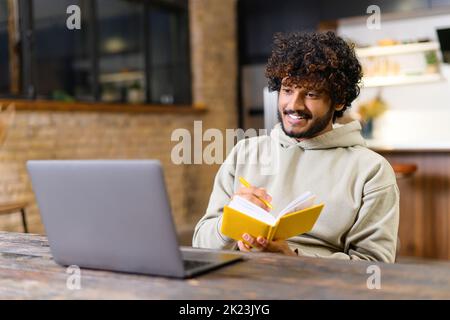 Handsome young man in smart casual wear is using laptop computer for watching online classes and webinars. He is taking notes at notebook, while sitting at contemporary home office Stock Photo