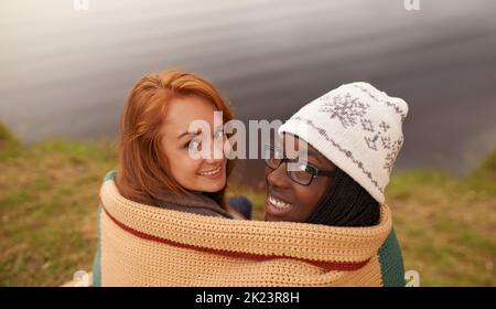 A cosy catch up by the lake. High angle portrait of two happy teenage girls huddled under a blanket beside a lake. Stock Photo