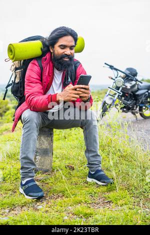 vertical shot of Happy smiling traveler busy using mobile phone while sitting on road side infrtont of motor bike - concept of technology, social Stock Photo