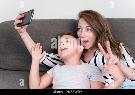 Cheerful mother holding smart phone making selfie with her little son showing victory sign. mom and kid boy sit on couch using gadget having fun taking self-portrait spend time together at home. Stock Photo