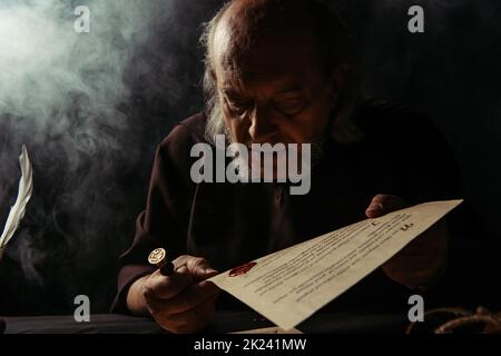 senior priest looking at wax seal on manuscript on dark background with smoke,stock image Stock Photo