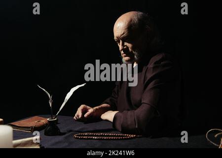 thoughtful monk sitting near inkpot and rosary at night isolated on black,stock image Stock Photo