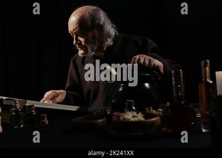 medieval wizard reading magic cookbook near pot and ingredients isolated on black,stock image Stock Photo