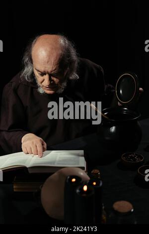 senior alchemist reading magic cookbook near pot isolated on black,stock image Stock Photo