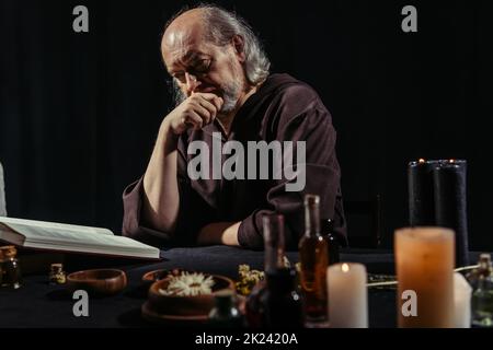 thoughtful alchemist reading magic cookbook near ingredients and candles isolated on black,stock image Stock Photo