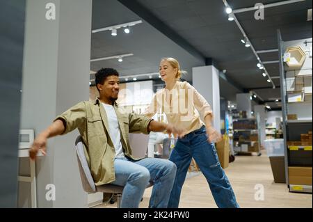 Happy funny married couple riding on office chair in furniture store having fun during shopping Stock Photo
