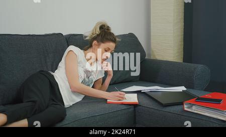 Pretty woman makes notes in notebook while lying on couch at home in a modern apartment. Is preparing for important meeting. Very focused prepares the Stock Photo