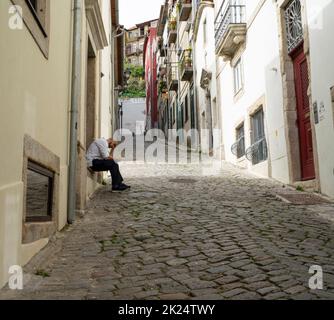 Porto, Portugal. March 2022.  A man rests sitting on the step of his house in a narrow street in the historic center of the city Stock Photo