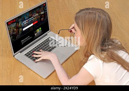 London, United Kingdom - January 13, 2022: Young woman on floor at home using laptop internet streaming service  browsing Netflix home page choosing m Stock Photo