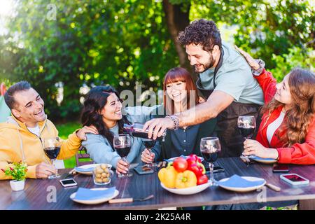 Man pouring red wine into glass while enjoying dinner party with friends outdoors in the backyard - happy friends having fun drinking wine in the terr Stock Photo