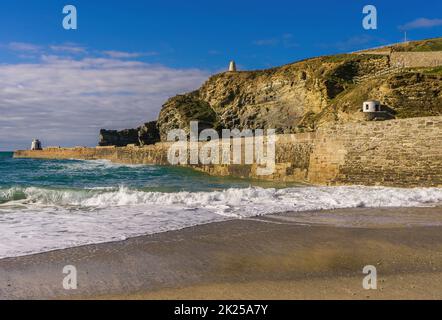 Waves break along the pier and onto the beach in Portreath, a small seaside village in Cornwall. On the pier and cliffs are the pilot's lookout towers. Stock Photo