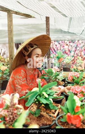 Asian woman with Vietnamese straw hat is smiling and holding potted flower in the garden. Stock Photo