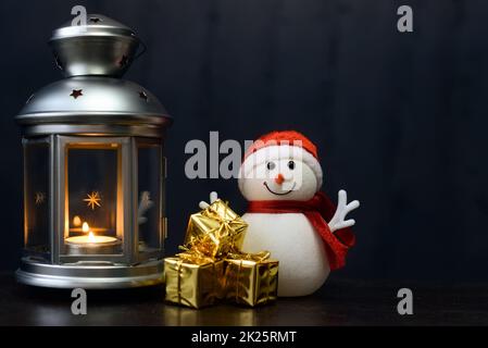 Christmas card: a snowman, a lantern with candles, bright shiny boxes with gifts on a dark background. Stock Photo