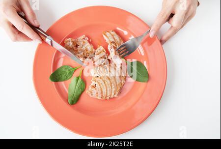 Close-up shot of a juicy delicious grilled tuna steak on a bright coral plate. Delicious and healthy food, proper nutrition. The girl is holding a fork and knife. View from above. Stock Photo