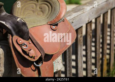 Old worn leather horse saddle on wooden fence, lit by sun. Stock Photo