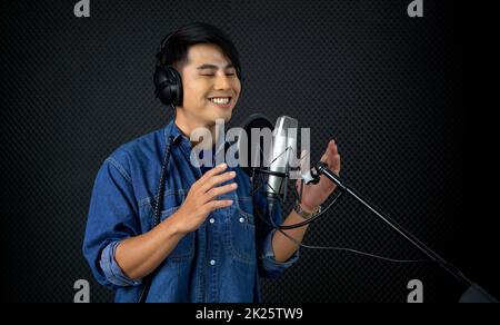 Young asian man with headphone singing in front of black soundproof wall. Musician producing music in professional recording studio. Stock Photo