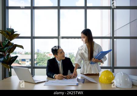 Asian project owner in suit and his assistant checking the construction drawing. Morning work atmosphere in a modern office. Stock Photo