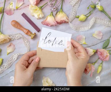 Hands with Card LOVE inside envelope near by pink flowers on a marble table Stock Photo