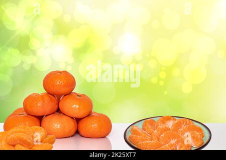 A pile of fresh ripe mandarines, citrus tangerines or orangen and slices of peeled tangerine on a plate over abstract bright background. Space for advertising. Clementines for health nutrition. Stock Photo