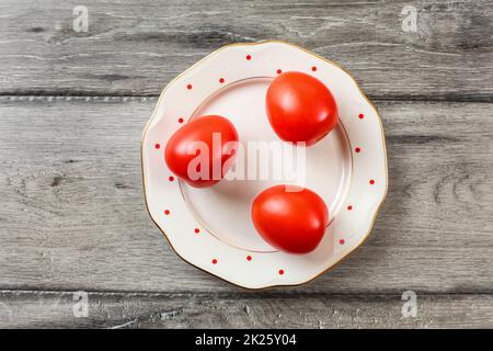 Three whole tomatoes on white plate with small red dots and golden rim, gray wooden desk, tabletop view. Stock Photo