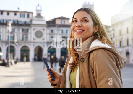 Copyspace photo of smiling young woman holding telephone with her hand wearing winter coat standing pensively looking up over city blurred background Stock Photo