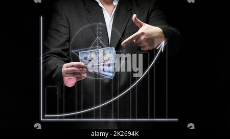 a businessman in a suit holds a pack of paper american dollars against the background of a holographic chart with growing indicators. The concept of high business profitability, income growth Stock Photo