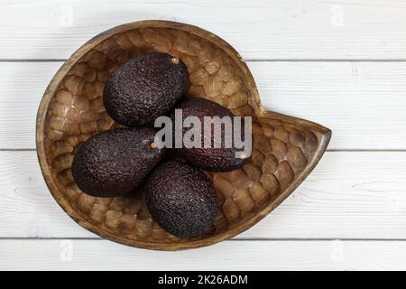 Four whole ripe brown avocados in wooden carved bowl on white boards desk. Tabletop view. Stock Photo