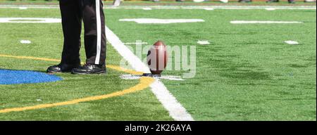 Football on the tee for kickoff of a high school game on a green turf field with thr referee standing next to the ball. Stock Photo