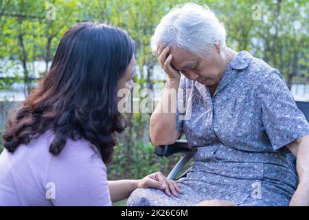 Caregiver help Asian senior or elderly old lady woman patient headache while sitting on wheelchair in park. Stock Photo