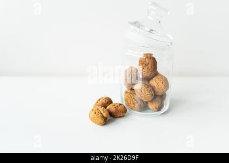 Beautiful whole walnut kernels in a glass jar, whole nuts in the shell can be seen nearby, on a white table. The concept of proper storage of walnuts. Selective focus. Stock Photo